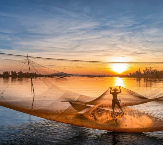 Silhouette of a person standing in a large, glowing fishing net on the water at sunset.