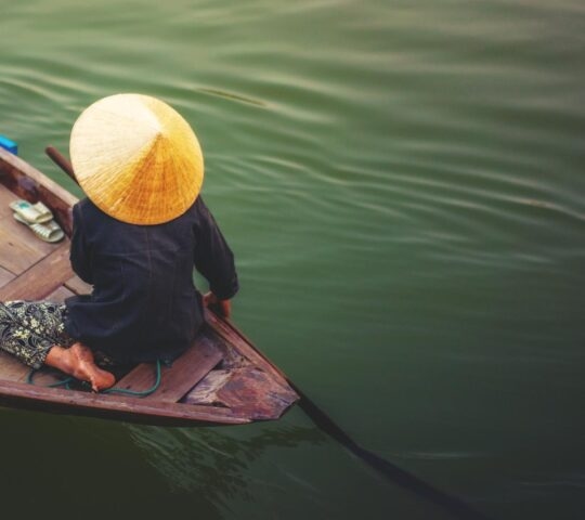 A person wearing a traditional conical hat rows a small wooden boat across dark, rippling water.