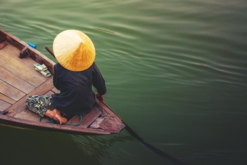 A person wearing a traditional conical hat rows a small wooden boat across dark, rippling water.
