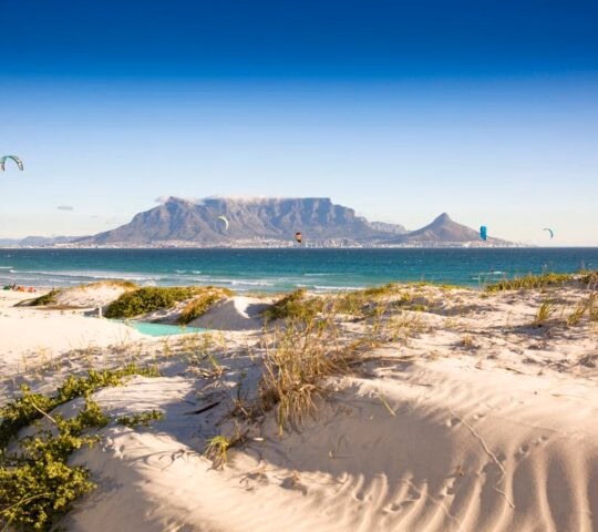 Sand dunes in the foreground overlook a blue bay with a large flat-topped mountain in the distant background.