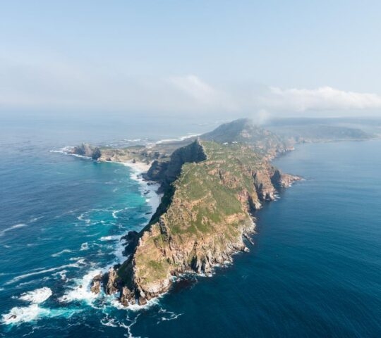 Aerial view of a long, narrow green peninsula surrounded by blue ocean water under a clear sky.