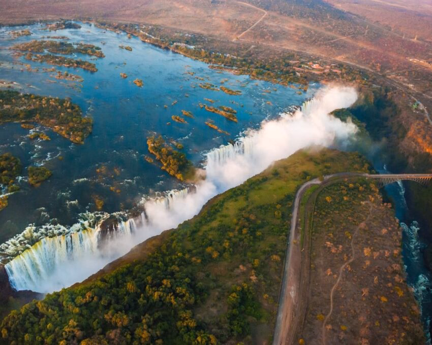 Aerial view of a wide waterfall with white mist rising into the air and lush green landscape along the riverbank.
