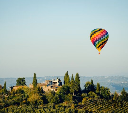 Colorful hot air balloon flying over a stone villa and rolling green vineyards under a clear sky.