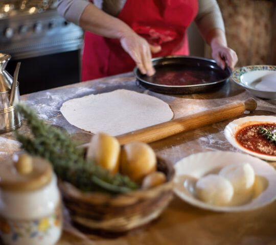 Close-up of hands preparing pizza dough on a wooden counter with sauce, cheese, and a rolling pin.