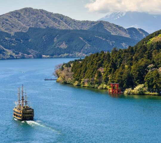 A large lake surrounded by green forested mountains, with a red Torii gate near the shore and a pirate ship sailing away.