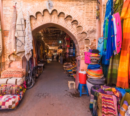 A stone archway at a market entrance surrounded by colorful hanging rugs, blankets, and stacks of cushions.