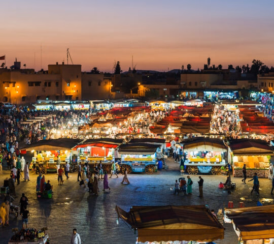 High angle view of a busy night market with many glowing white and red stalls crowded with people at dusk.
