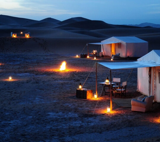A desert camp at night with glowing lanterns and a small fire outside white tents against dark sand dunes.