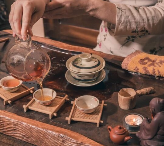 A close-up of hands pouring dark tea from a glass pitcher into small ceramic cups on a wooden tray, with a burning candle.