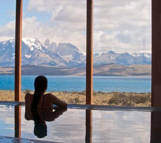 Silhouette of a person in a pool looking out large windows at a turquoise lake and snow-covered mountains.