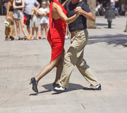 A woman in a red dress and a man in tan pants dancing tango in an open paved square.