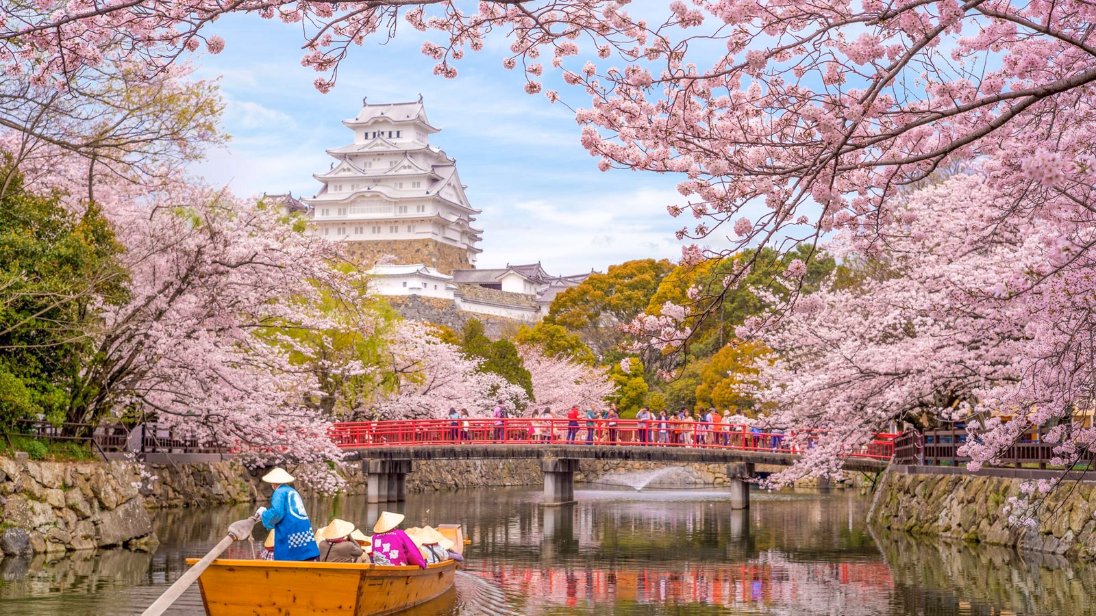A white Japanese castle framed by cherry blossoms with a red bridge crossing a moat and a small boat.