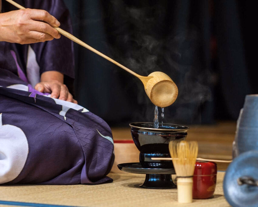 Close-up of a tea ceremony showing water poured from a wooden ladle into a black bowl on a tatami mat.