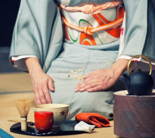 A seated person in a kimono with a tea whisk, red container, and black teapot on a tatami floor.