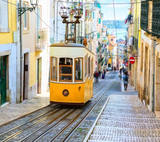 A yellow funicular tram travels down a steep street between tall, colorful buildings toward the sea in the distance.
