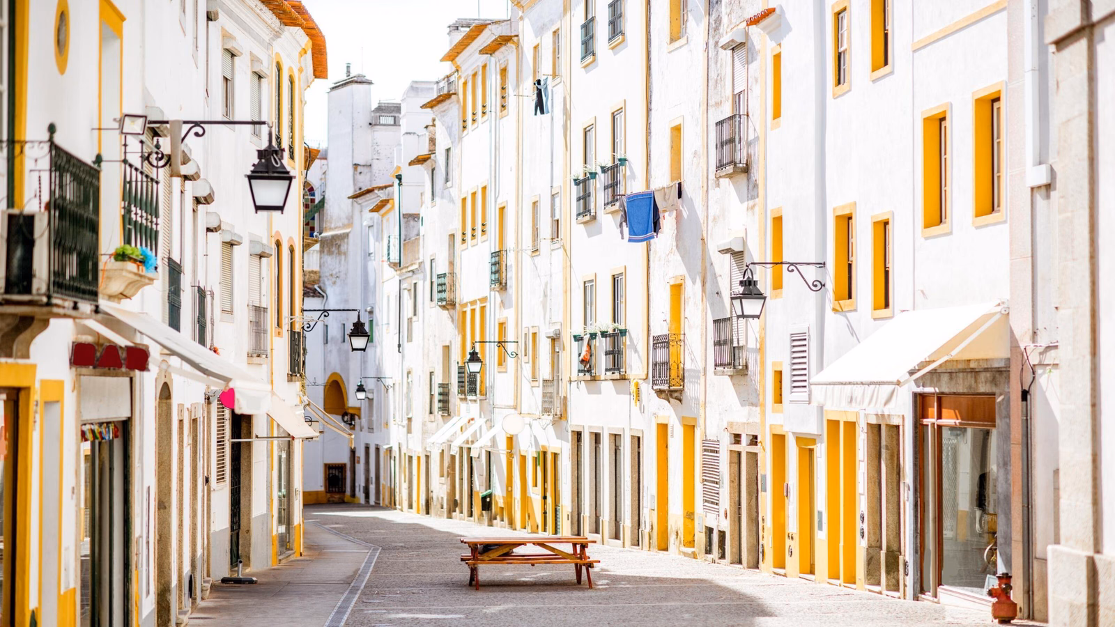 Narrow sunlit cobblestone street with tall white and yellow buildings and hanging street lanterns.