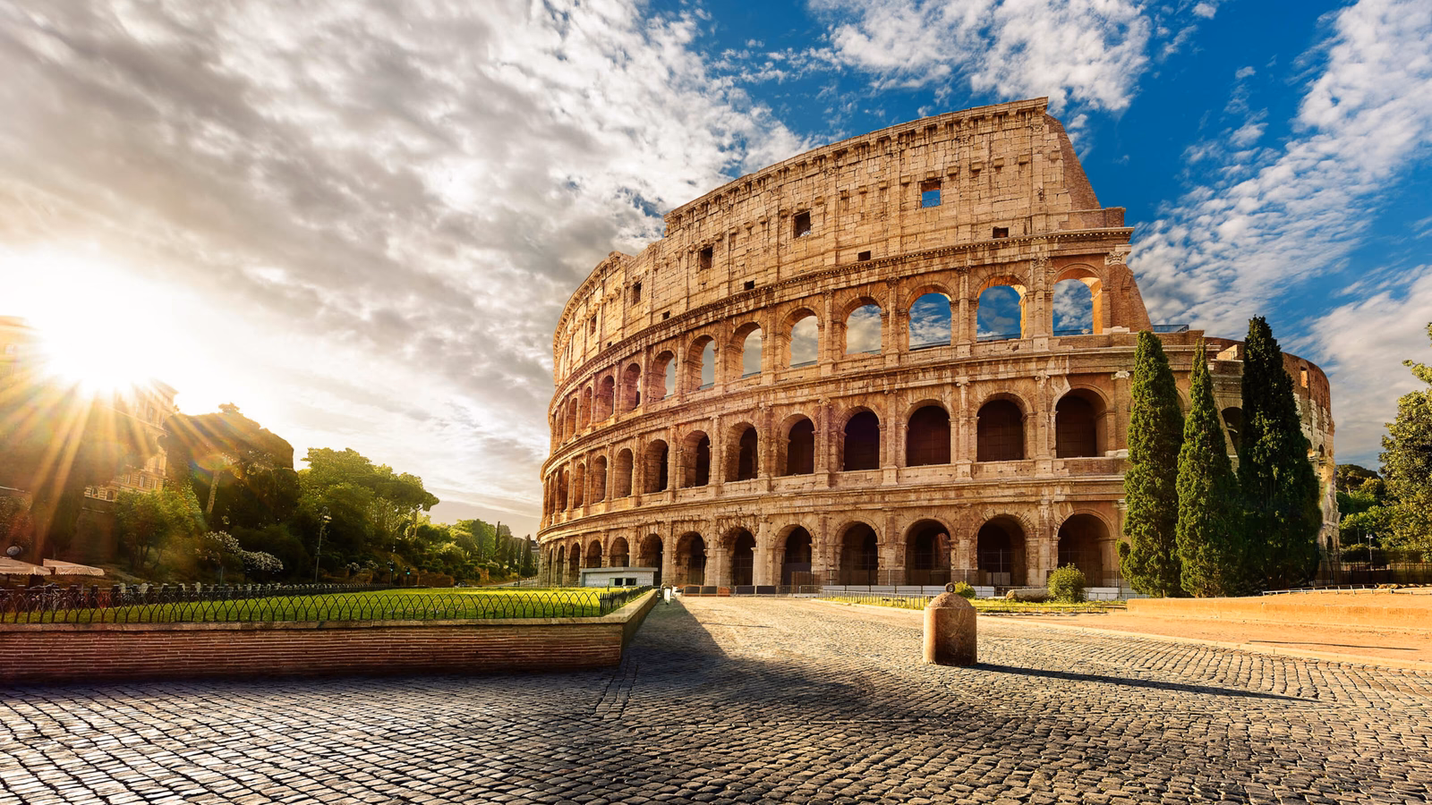 The sun shining through the side of the ancient Roman Colosseum amphitheater with a blue, cloudy sky above.