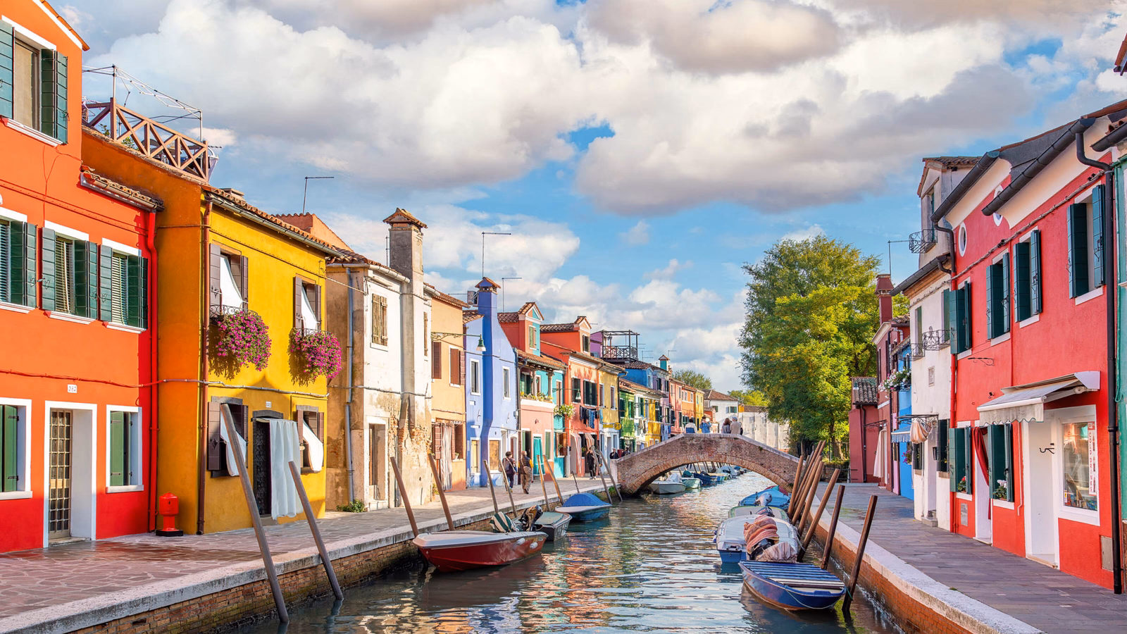 Vibrant multi-colored houses reflected in a calm canal with a small footbridge and moored boats.