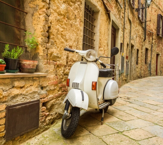 A white vintage scooter parked in a narrow cobblestone alleyway against an old stone wall with plants.