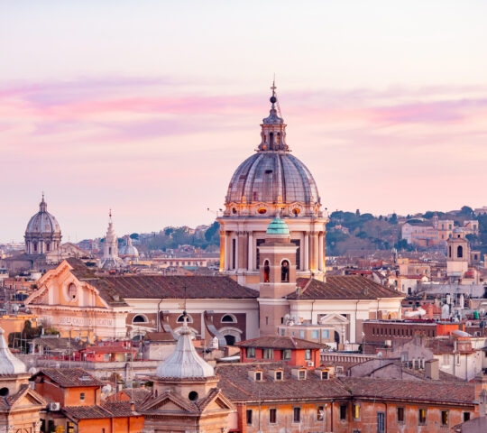 Aerial view of Rome skyline at sunset with large domes and historic buildings under a colorful sky.