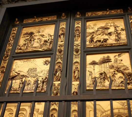 Close-up of golden bronze relief panels on a large door featuring intricate carvings of figures and landscapes.
