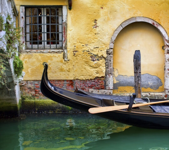Close-up of a black gondola on green water next to a textured yellow wall and a stone bridge in Venice.