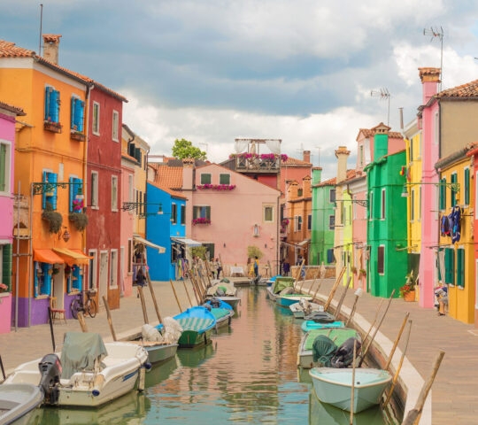 A row of vibrant, colorful houses along a canal with moored boats under a cloudy sky in Burano.