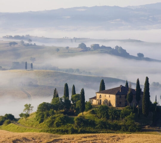 A stone house on a hill is surrounded by cypress trees and low-lying white fog in a valley.