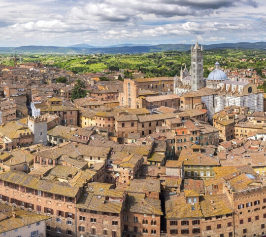Aerial view of Siena, Italy, showing dense terracotta roofed buildings and the cathedral tower under a cloudy sky.