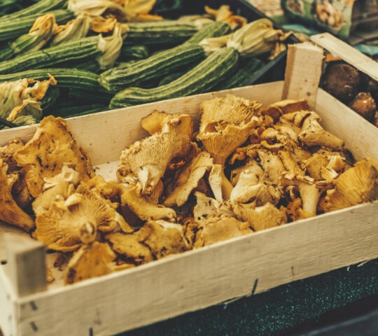 A wooden crate filled with golden chanterelle mushrooms sits next to a pile of green zucchini at an outdoor market.