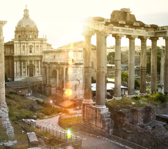 Sunlight flares over ancient stone ruins and columns of the Roman Forum under a bright sky.