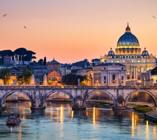 St. Peter's Basilica and a stone bridge over the Tiber River illuminated at twilight under an orange sky.