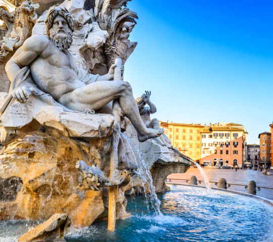 Elaborate stone fountain sculpture of a seated man with water flowing into a basin in a city square.