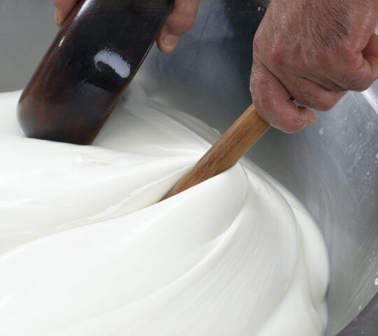 Close-up of hands using a wooden stick to stir a thick, white, creamy substance in a metal container.