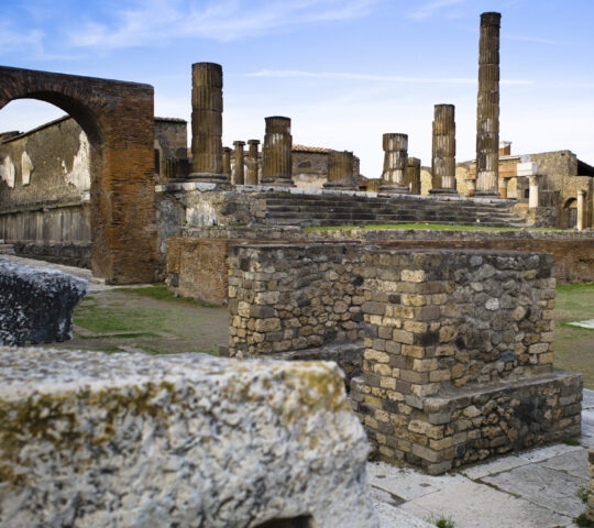 Historic ruins with stone pillars, brick foundations, and a large archway under a bright sky.