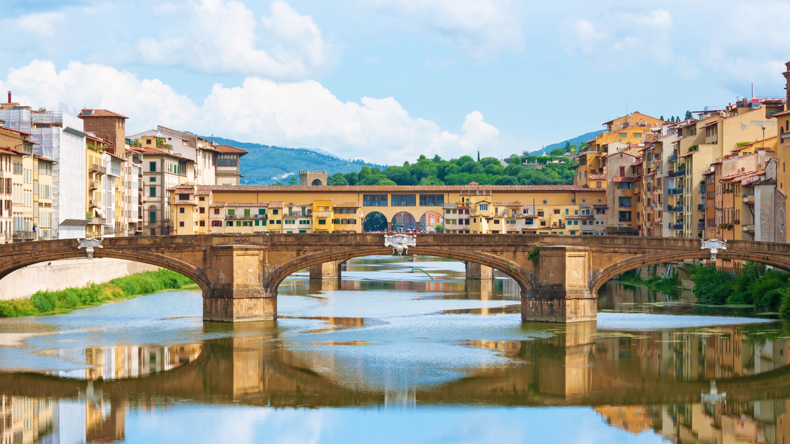 A stone bridge with buildings built upon it reflects in the calm river water under a cloudy sky.