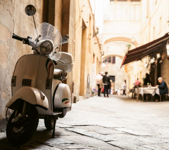 A white vintage scooter parked on a cobblestone street in a narrow, sunlit alley with an outdoor cafe.