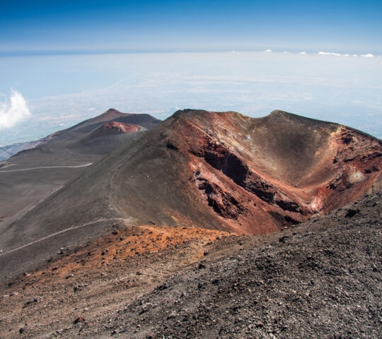 A wide view of a large, barren volcanic crater with dark rocky soil and a clear blue sky above.
