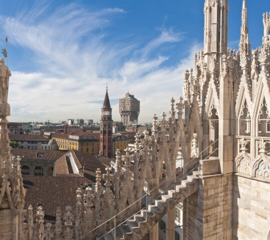 View from a stone rooftop showing intricate Gothic spires and pinnacles overlooking a city under a blue, wispy sky.
