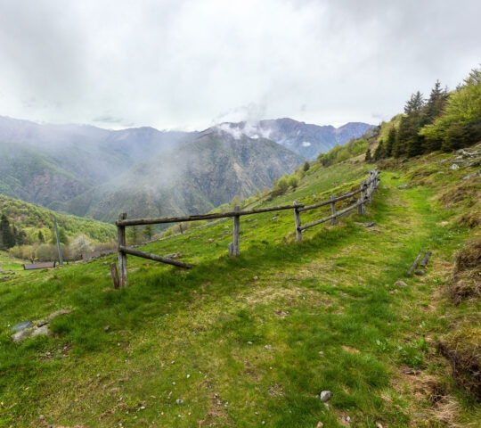 A simple wooden fence runs along a grassy hillside overlooking a misty mountain valley under a cloudy sky.
