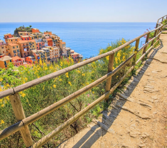 A narrow dirt path with a wooden railing overlooks a colorful hillside village and the vast, blue Mediterranean Sea.