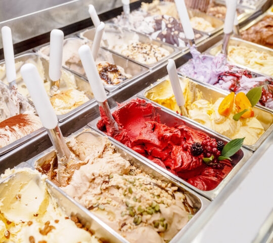 Multiple metal tubs of colorful Italian gelato, including red berry and pale green flavors, inside a shop freezer.