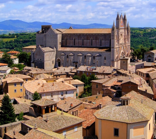 A large cathedral with a pointed facade stands high above the orange roofs of a surrounding Italian village.