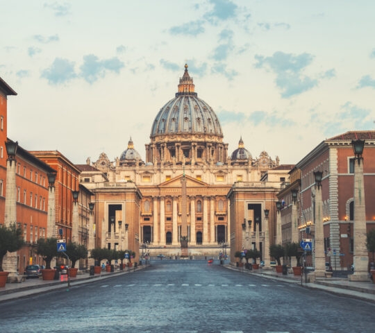 A centered view of a long city street leading to the large dome of St. Peter's Basilica under a pale sky.