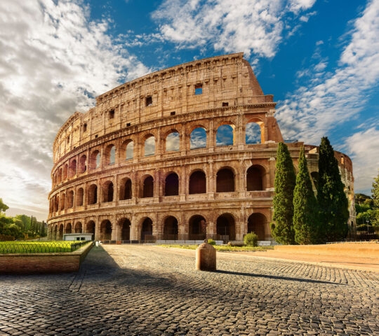The historic stone arches of the Roman Colosseum under a blue sky with scattered white clouds.