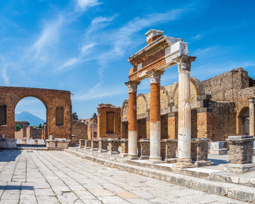 Ruined stone columns and an archway of an ancient Roman structure at Pompeii with mountains in the background.