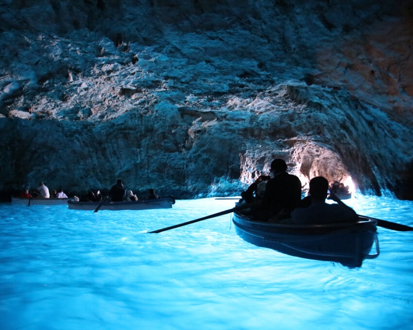 Tourists in small boats inside a dark sea cave where the water is glowing a vibrant electric blue.