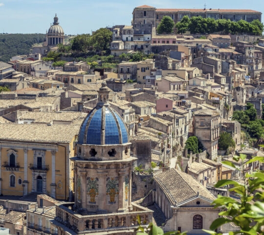 A wide view of a sun-drenched Italian hilltop town featuring a blue-domed church and many stone houses.