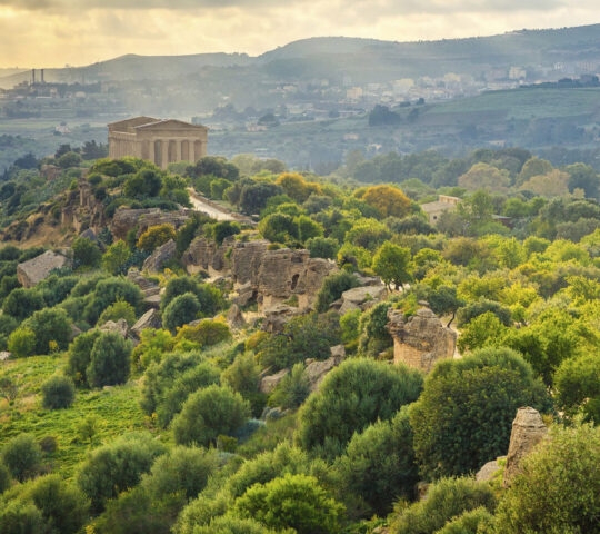 A distant view of a preserved ancient temple on a hill surrounded by green trees and rocky terrain.