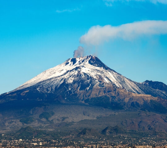 Snow-covered peak of Mount Etna volcano with smoke emitting from the crater against a clear blue sky.